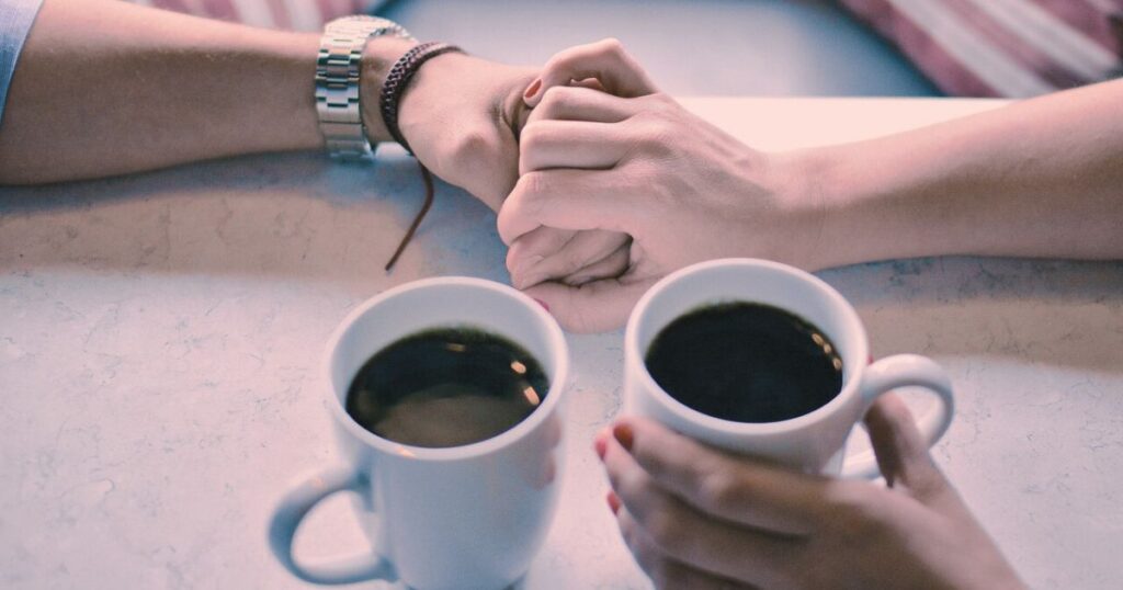 カフェで向かい合いながら手をつなぐ男女の手元。言葉がなくてもつながりを感じる静かなデートの瞬間。