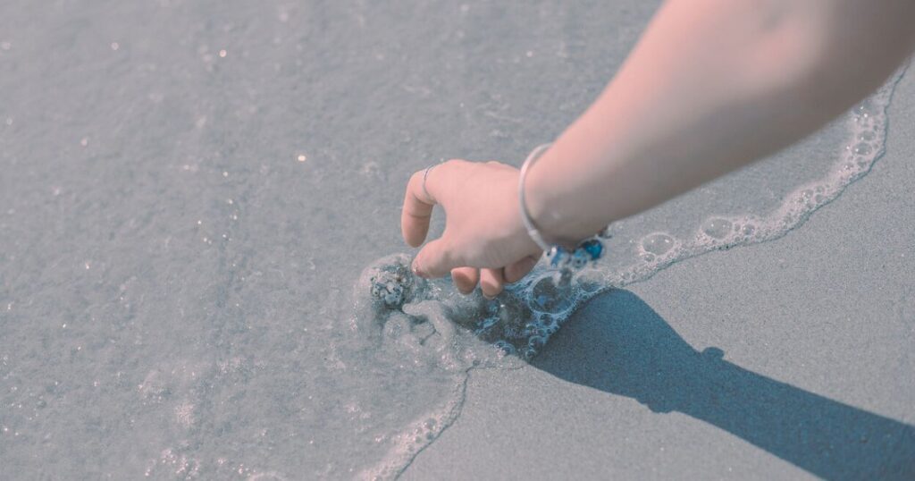 波にそっと手を伸ばす女性の手元。柔らかな光の中で静かな時間を感じる様子。