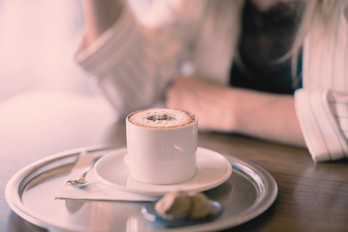 淡い光の中で、テーブルに置かれた温かいコーヒーを前に、女性がそっと手を添えている写真。落ち着いた時間が流れる雰囲気が伝わる。